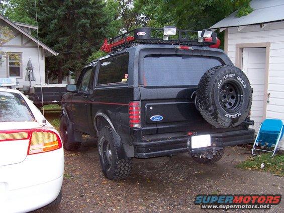 Black 1996 Ford Bronco parked in a driveway, spare tire on back, roof rack with lights