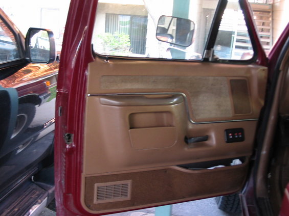 Red car door interior with tan panel, armrest, and speaker grille. A dark vehicle is reflected in the side mirror