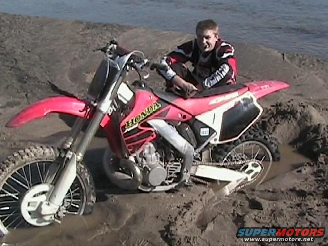 Guy sitting on a red Honda dirt bike stuck in the mud near water