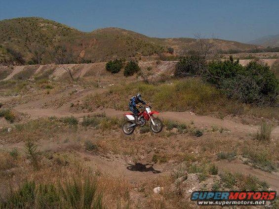 Red dirt bike mid-air, rider in blue and black gear, dusty terrain, sunny day