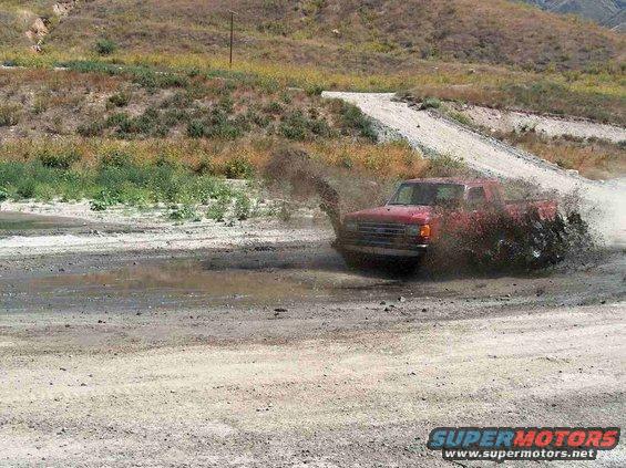 Red pickup truck kicking up mud while driving through a puddle. The truck appears to be from the late 1980s