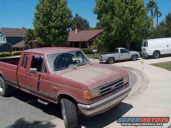 Red Ford F-series pickup from the late 80s parked on a residential street. A silver pickup and white van are behind it