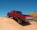 1989 Ford F-250 - This red 1989 Ford F-250 pickup truck is captured in a rear 3/4 view as it navigates a dirt incline under a clear blue sky.