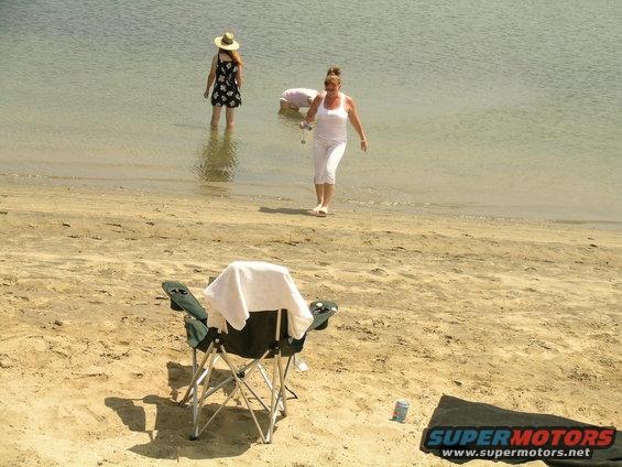 Two women and a child wading in shallow water at a sandy beach, with a folding chair and towel in the foreground