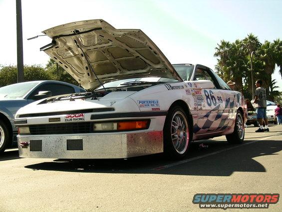 White race car with hood up, number 86 on door, checkered flag pattern on side. Another car parked next to it