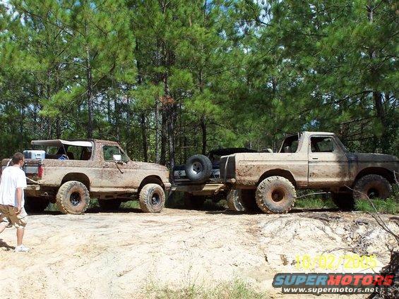 Two muddy 1980s Ford Broncos are parked on a dirt trail with trees in the background