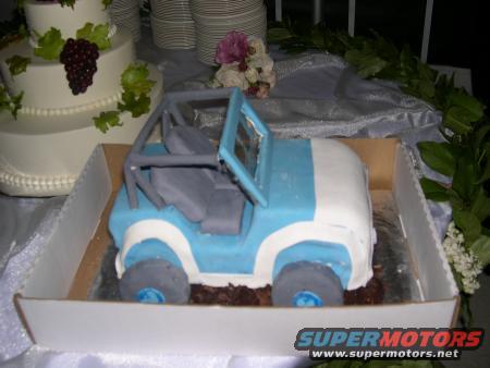 Cake decorated to look like a blue and white jeep, with a wedding cake in the background
