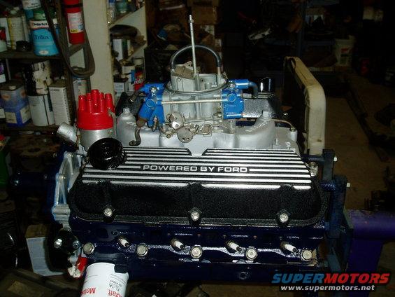 Ford V8 engine with black valve covers reading "POWERED BY FORD", silver intake manifold, and red distributor cap