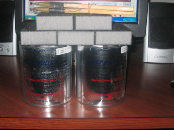 Two cans of automotive polish sit on a desk, topped with gray foam blocks. A computer monitor and speakers are in the background