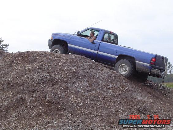 Blue pickup truck with a man inside climbing a dirt mound. The truck has a single cab and a long bed