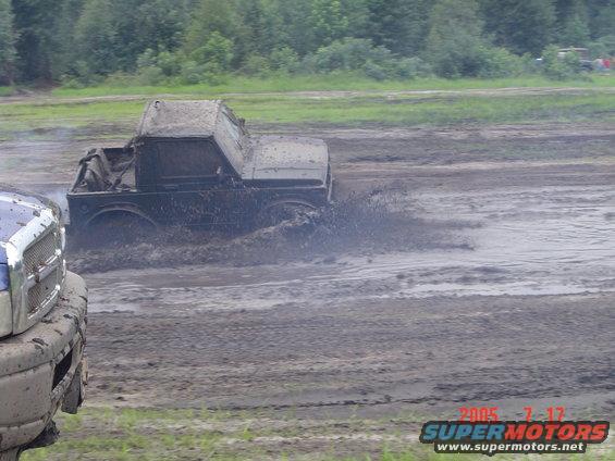 Muddy truck splashing through a bog. Another vehicle is partially visible on the left