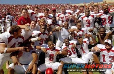 Football team in white and red uniforms celebrating on a sunny day with a stadium crowd in the background
