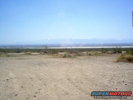 Desert landscape with dry brush, dirt ground, and distant mountains under a clear blue sky. Supermotors.net logo in lower right