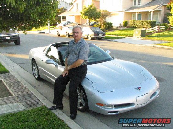 Silver convertible sports car parked on street, man sitting on hood. Another car and truck visible in background