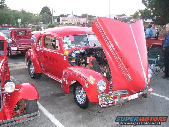 Red hot rod coupe with hood open, showing engine. Another red vehicle is parked behind it