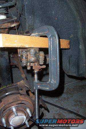 Brake caliper compressed by C-clamp and wood block, with wheel hub visible