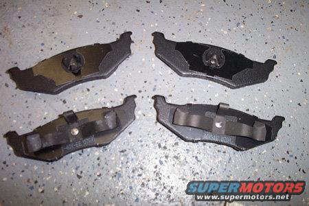 Four brake pads laid out on a speckled gray floor. Some have metal clips attached