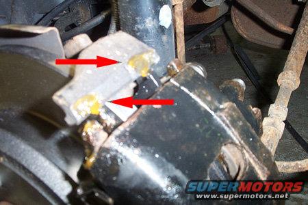 Close-up of a vehicle's disk brake caliper with red arrows pointing to yellow grease
