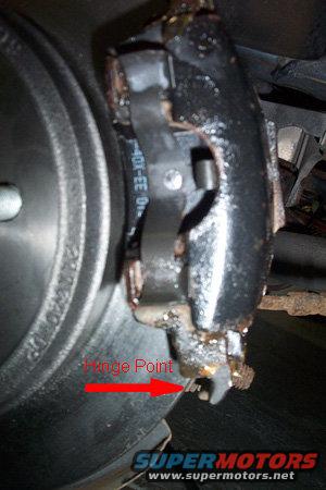 Close-up of a dirty brake caliper and rotor with a red arrow pointing to a hinge point