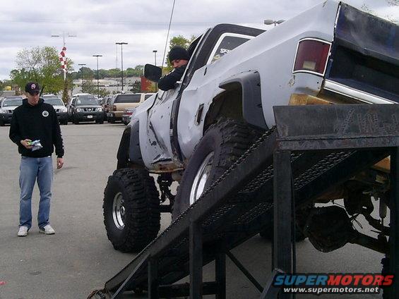 Gray lifted pickup truck with large tires on a metal ramp, driver visible, another person standing nearby