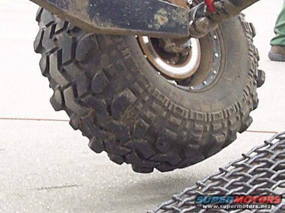 Close-up of a large off-road tire with aggressive tread, attached to a vehicle's suspension. Metal grating is visible in the foreground