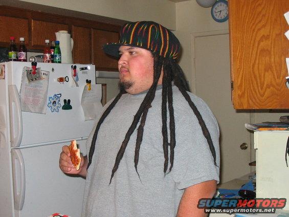 Man with dreadlocks and a colorful hat holds a slice of pizza in a kitchen