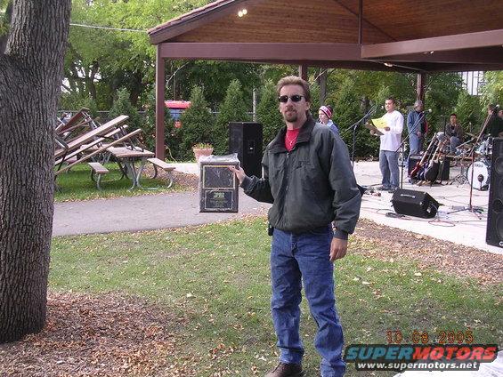 Man in jeans and jacket holds a plaque with text and a logo; outdoor event with band setup in background