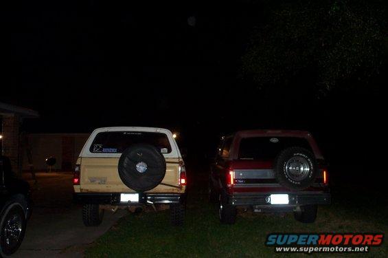 Two SUVs parked at night, rear view. The one on the left is tan with a spare tire on the back. The one on the right is red with a spare tire on the back