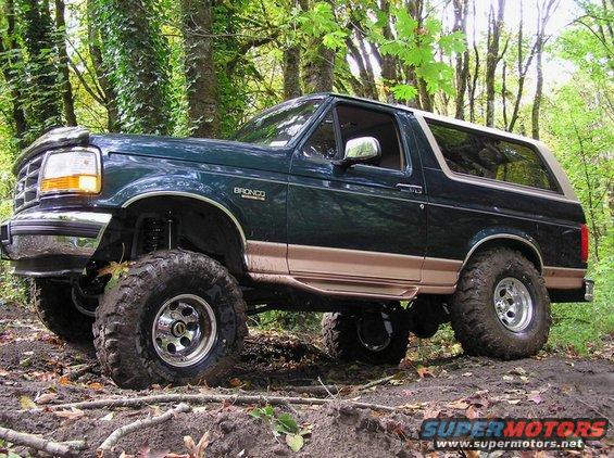 Lifted dark green 1995 Ford Bronco with tan trim, off-roading on a dirt path in the woods