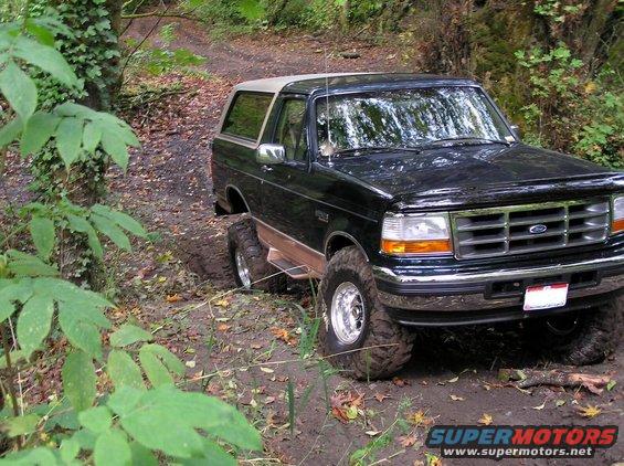 Black Ford Bronco with tan top on a dirt trail, front right tire stuck in mud