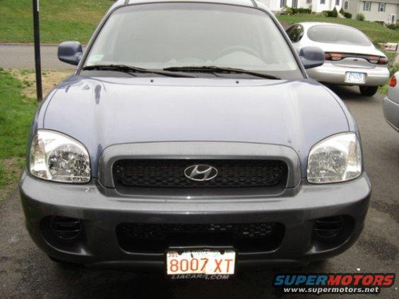 Front view of a blue Hyundai Santa Fe with a dark gray grille and bumper. A silver Ford Taurus is parked behind it