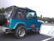1979 Ford Bronco - A teal Jeep Wrangler drives down the road with its soft top up and a spare tire mounted on the back.