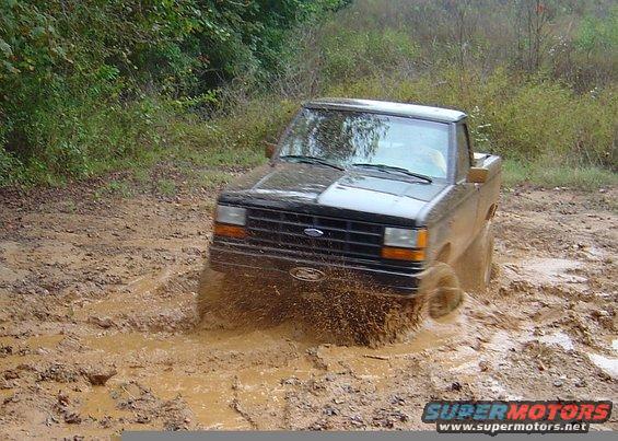 Black pickup truck with a dented front fender driving through deep mud, splashing water
