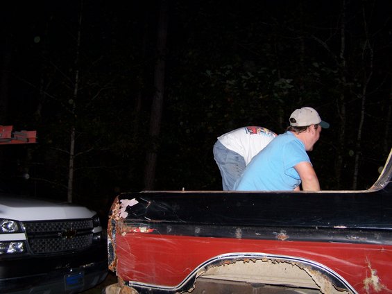 Two guys are working on the rusty bed of a red and black pickup truck. Another white truck is parked next to it