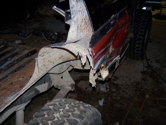 Heavily rusted and damaged rear quarter panel and fender of a truck, showing exposed metal and red paint