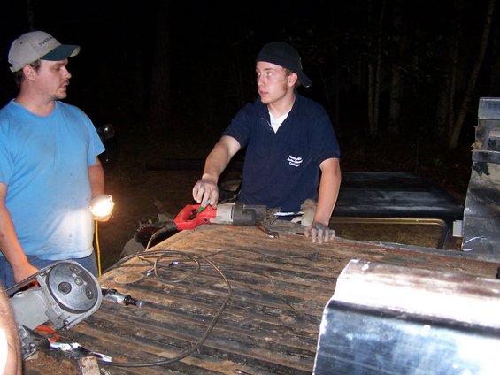 Two men work on a dark, wooden truck bed at night with tools. One man holds a flashlight