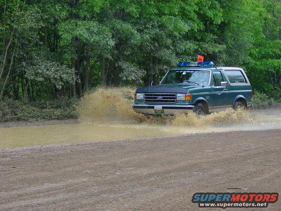 Green 1989 Ford Bronco splashing through a muddy puddle on a dirt road, trees in background