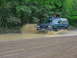 1989 Ford Bronco - 1989 Ford Bronco navigates a muddy obstacle on a dirt path surrounded by trees.