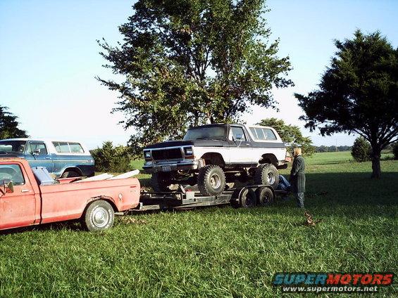 Two-tone Ford Bronco on a trailer, lifted with large tires. A red pickup truck tows the trailer. A blue SUV is parked nearby