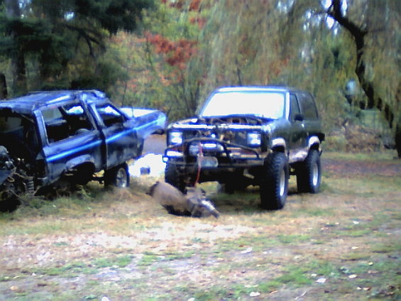 Two off-road vehicles, one dark green with hood open and a transmission on the ground in front. A blue pickup truck is to the left