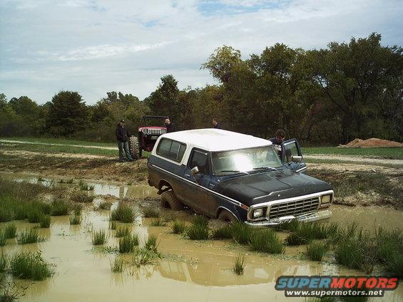 Dark blue and white SUV stuck in mud, front end submerged. A red Jeep is behind it with two people standing nearby