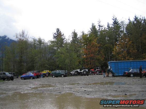Several off-road vehicles parked in a muddy lot with trees and a blue shipping container