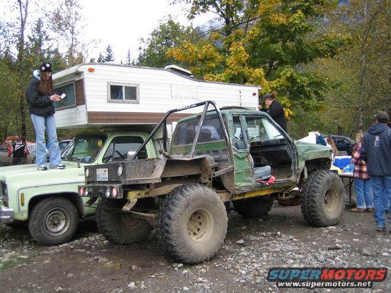 Modified green truck with large tires and roll cage, parked next to a pickup with a camper shell. People are standing around