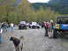 1990 Ford Bronco - Several off-road vehicles are parked in a gravel lot surrounded by trees, with people and a dog present.