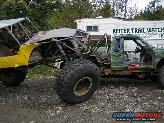 Custom yellow buggy with large tires next to a modified green pickup truck. Trailer in background reads 'REITER TRAIL WATCH'