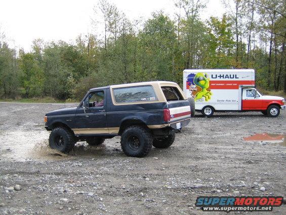 Dark blue Ford Bronco with tan trim kicking up mud, U-Haul truck behind it