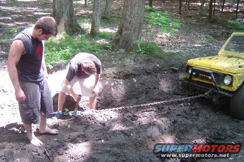 Yellow 1987 Suzuki Samurai stuck in mud, front view, with two men attempting to pull it out with a chain