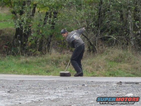 Man sweeping debris from a paved road in a rural area with trees in the background