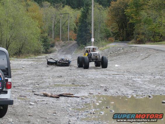 Yellow buggy with big tires tows a pile of debris on a dirt road. White Jeep visible on left
