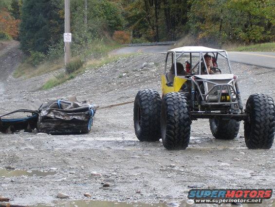 Yellow off-road buggy with large tires tows a crushed car on a dirt road